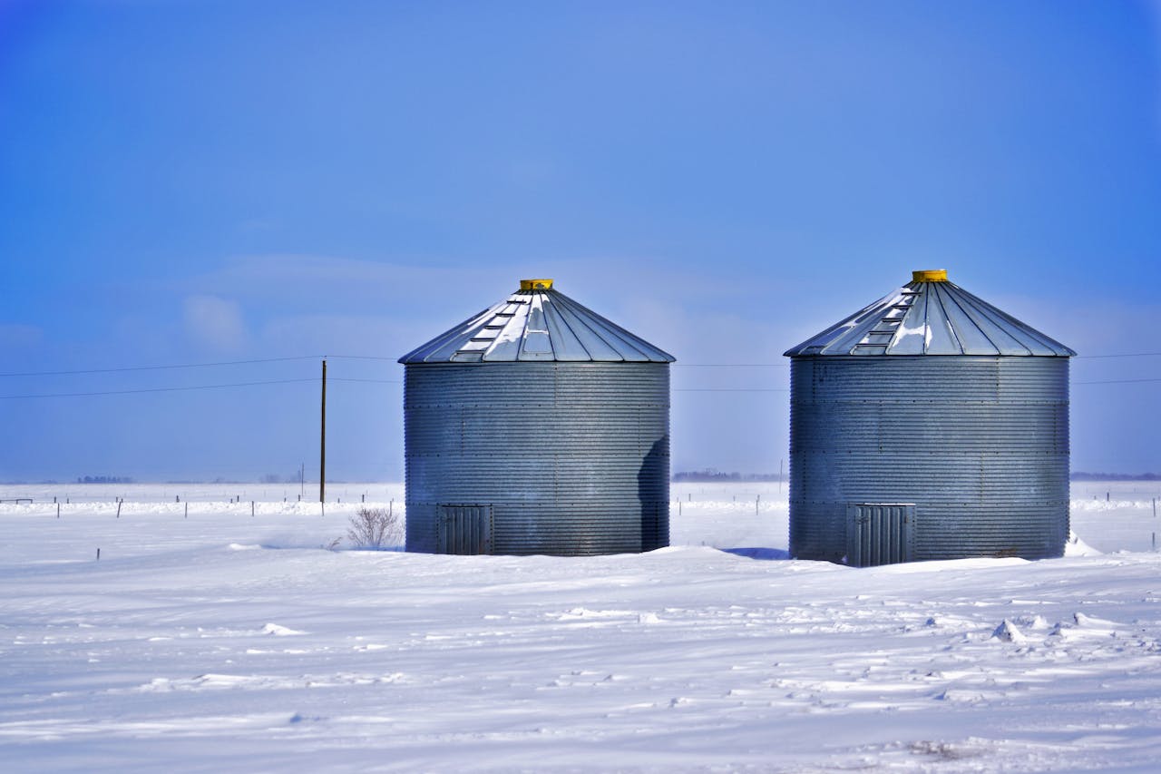 Two steel silos stand isolated in a snowy landscape under a clear blue winter sky.